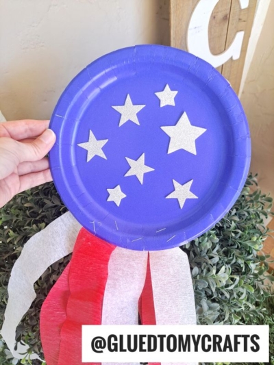 A hand holds a blue paper plate flag decorated with white stars and red and white crepe paper streamers hanging from the bottom. The plate is held near green foliage. Text reads @GLUEDTOMYCRAFTS.