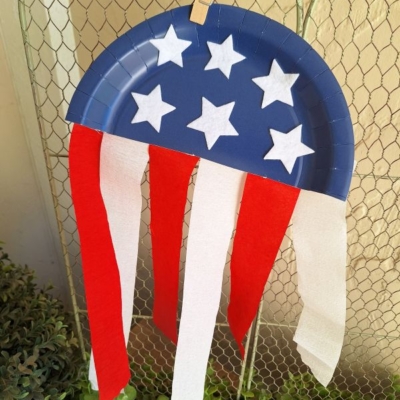 A blue paper plate half decorated with white stars and hanging red and white streamers, clipped to a wire fence. The text @GLUEDTOMYCRAFTS appears at the bottom.