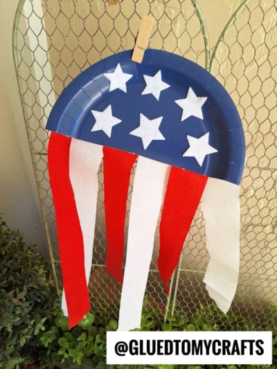 A blue paper plate half decorated with white stars and hanging red and white streamers, clipped to a wire fence. The text @GLUEDTOMYCRAFTS appears at the bottom.
