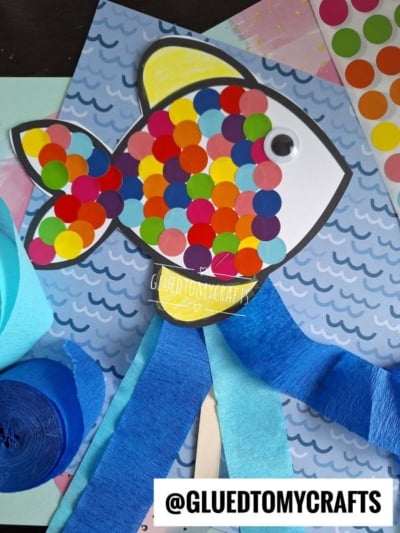 A colorful paper fish decorated with multicolored dot stickers and a googly eye is placed on a blue wavy background. Strips of blue crepe paper hang below, resembling water. @GLUEDTOMYCRAFTS is written at the bottom.