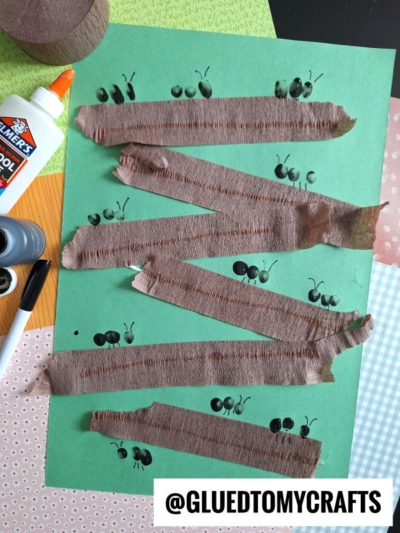 A green sheet of paper with zigzag strips of brown crepe paper glued across it, black ant drawings in between, a bottle of glue, a marker, and scissors nearby. The username @GLUEDTOMYCRAFTS is at the bottom.