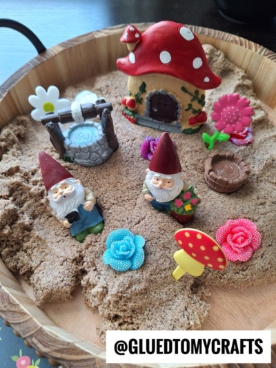 A miniature fairy garden scene in a round tray with sand, featuring two gnome figurines, a mushroom house, a well, and colorful artificial flowers. @GLUEDTOMYCRAFTS is labeled at the bottom.