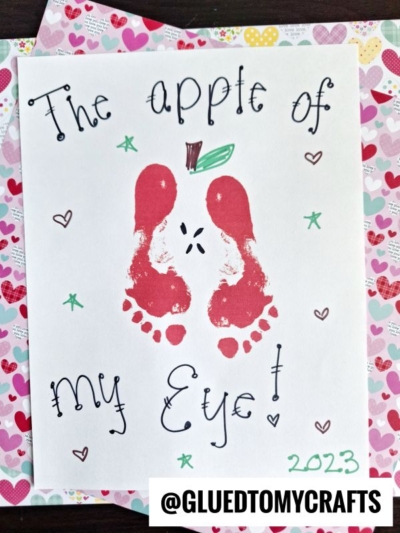 A handmade card with red painted footprints forming an apple, green leaves at the top, and The apple of my eye! in playful lettering. Small hearts, 2023, and a HANDPRINT FATHER'S DAY theme make it extra special.