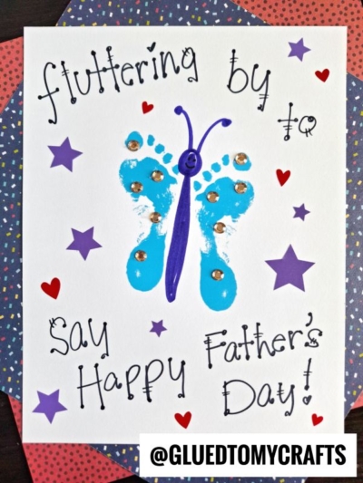 Handmade HANDPRINT FATHER'S DAY card featuring a blue butterfly crafted from painted footprints, adorned with rhinestones and surrounded by hearts and stars. Text reads: Fluttering by to say Happy Fathers Day!.
