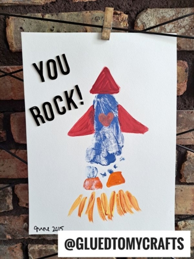 A child’s painted rocket ship with a red heart in the center, orange flames below, and “YOU ROCK!” in bold letters above. Perfect for a HANDPRINT FATHER'S DAY craft. The art is clipped to a wire against a brick background. Text: @GLUEDTOMYCRAFTS.