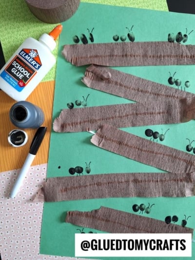 A craft project with green paper, brown crepe paper zigzagged and glued on, black ant stamps, a bottle of Elmer’s glue, black ink, and a marker. @GLUEDTOMYCRAFTS is printed at the bottom.
