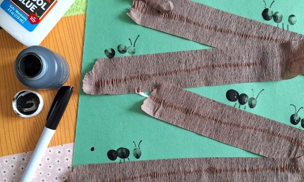 A craft project with green paper, brown crepe paper zigzagged and glued on, black ant stamps, a bottle of Elmer’s glue, black ink, and a marker. @GLUEDTOMYCRAFTS is printed at the bottom.