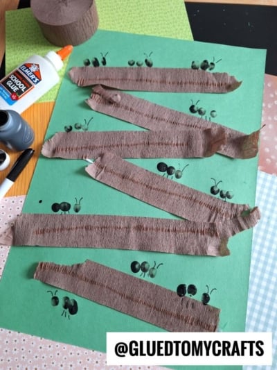 A green paper with black ant fingerprints and zigzag strips of brown crepe paper, surrounded by glue, marker, and patterned paper. The handle @GLUEDTOMYCRAFTS is at the bottom right.
