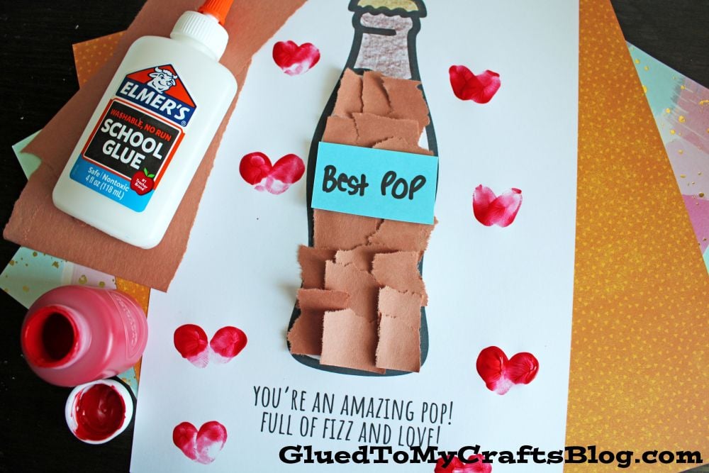 A handmade craft features a soda bottle with “Best Pop” on it, made from torn brown paper on white paper. Surrounding are red paint hearts, a glue bottle, and colored paper. Text reads: “You’re an amazing pop! Full of fizz and love!”.