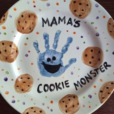 A decorated plate with chocolate chip cookies painted around the edges, a blue handprint in the center with a smiling face to resemble Cookie Monster, and the words MAMAS COOKIE MONSTER—perfect for adorable Mother's Day keepsakes.