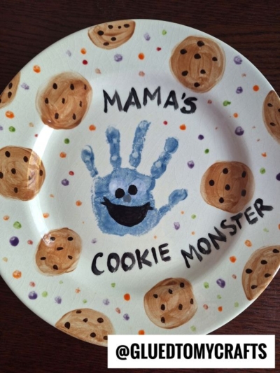 A decorated plate with chocolate chip cookies painted around the edges, a blue handprint in the center with a smiling face to resemble Cookie Monster, and the words MAMAS COOKIE MONSTER—perfect for adorable Mother's Day keepsakes.