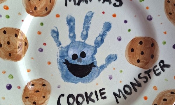 A decorated plate with chocolate chip cookies painted around the edges, a blue handprint in the center with a smiling face to resemble Cookie Monster, and the words MAMAS COOKIE MONSTER—perfect for adorable Mother's Day keepsakes.