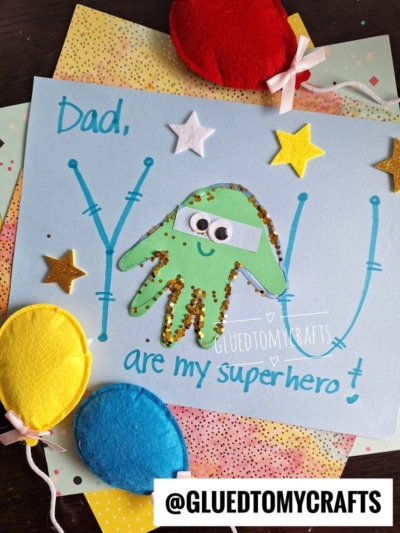 A handmade HANDPRINT FATHER'S DAY card features Dad, you are my superhero! with a green handprint octopus decorated with googly eyes and glitter, surrounded by star stickers and colorful paper balloons.