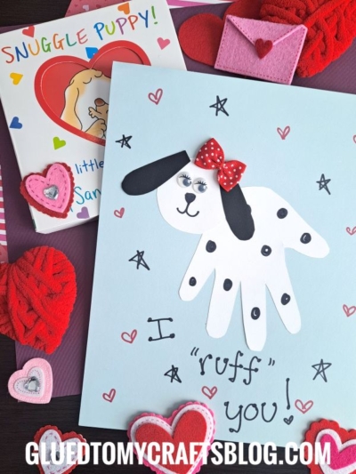 A handmade HANDPRINT FATHER'S DAY dog card, crafted with a white handprint, black spots, googly eyes, and a red bow, sits on a blue page with felt hearts. The text says “I ‘ruff’ you!” A book titled “Snuggle Puppy!” peeks out nearby.