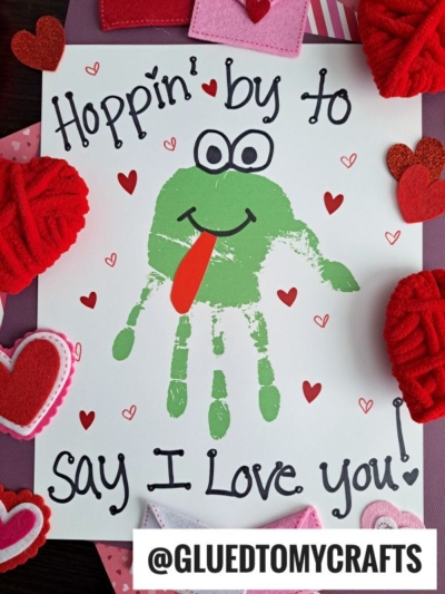 A handmade Father's Day card features a green frog made from a handprint, with a red tongue and googly eyes. The text says Hoppin by to say I love you! Decorated with red and pink hearts.