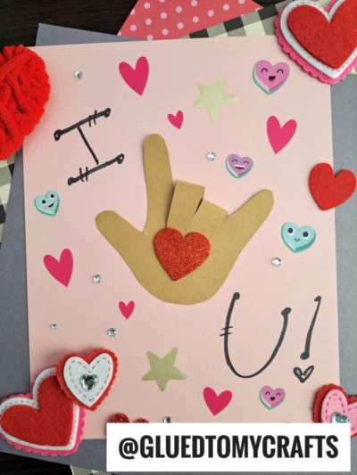 A pink card decorated with paper hearts, rhinestones, and a HANDPRINT FATHER'S DAY cutout forming the ASL I love you sign, with U! written in marker and @GLUEDTOMYCRAFTS printed at the bottom.