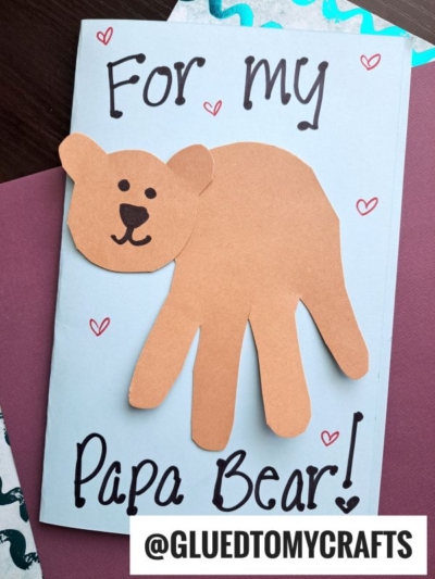 A handmade HANDPRINT FATHER'S DAY card featuring a brown paper bear with a handprint body and "For my Papa Bear!" on a light blue background, decorated with small red hearts.