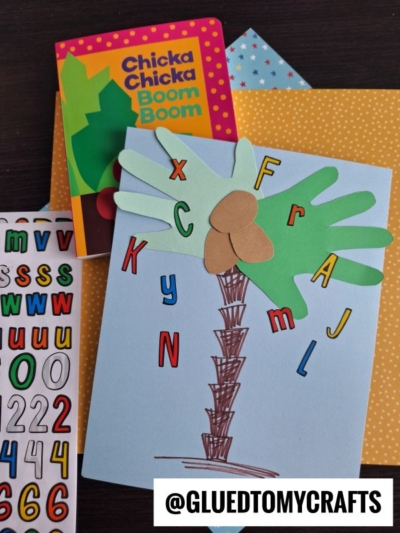 A handmade card inspired by “Chicka Chicka Boom Boom” features a paper palm tree with colorful letter cutouts, a book, number stickers, and yellow polka-dot paper in the background. A watermark reads @GLUEDTOMYCRAFTS.