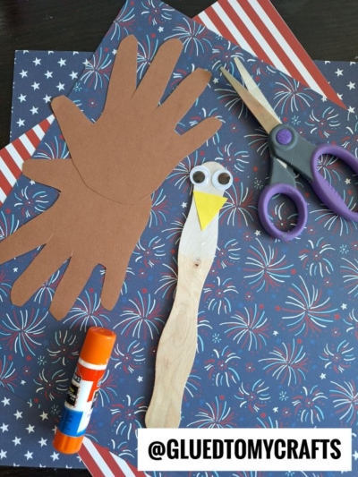 A craft project featuring hand-shaped brown paper cutouts, a popsicle stick decorated as an eagle with googly eyes and a yellow beak, scissors, glue stick, and patriotic-themed paper.