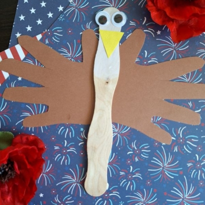 A turkey craft made from a wooden stick, brown paper handprints for wings, googly eyes, and a yellow paper beak is displayed on patriotic star-patterned paper with red faux flowers.