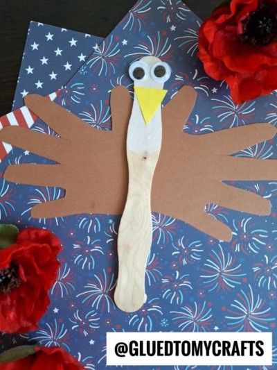 A turkey craft made from a wooden stick, brown paper handprints for wings, googly eyes, and a yellow paper beak is displayed on patriotic star-patterned paper with red faux flowers.