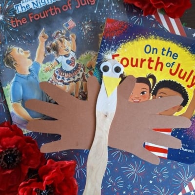 A craft eagle made from a popsicle stick and brown paper hand cutouts is placed on top of two Fourth of July-themed children’s books, surrounded by red artificial flowers. @GLUEDTOMYCRAFTS is printed at the bottom.