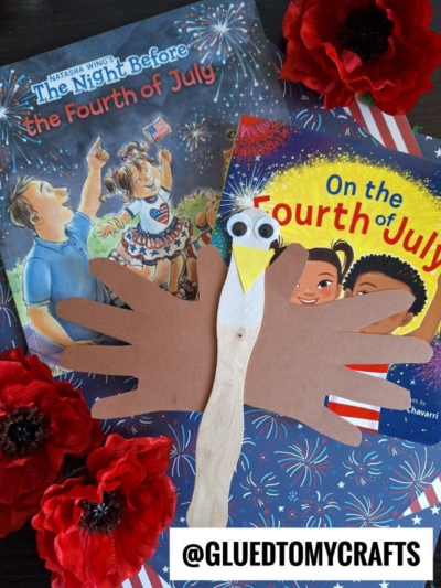 A craft eagle made from a popsicle stick and brown paper hand cutouts is placed on top of two Fourth of July-themed children’s books, surrounded by red artificial flowers. @GLUEDTOMYCRAFTS is printed at the bottom.
