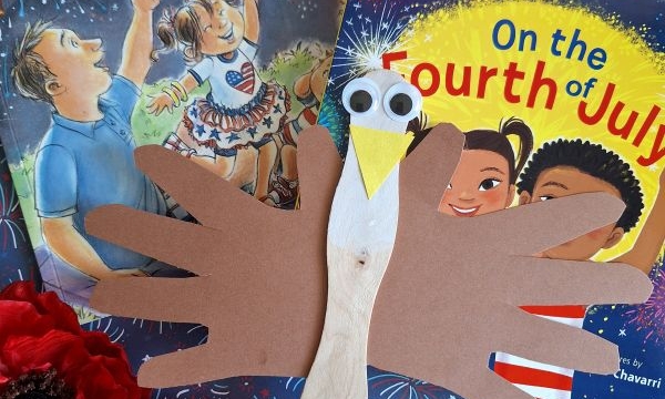A craft eagle made from a popsicle stick and brown paper hand cutouts is placed on top of two Fourth of July-themed children’s books, surrounded by red artificial flowers. @GLUEDTOMYCRAFTS is printed at the bottom.