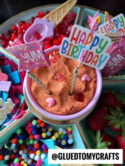 A colorful DIY birthday play kit with a bowl of orange play dough topped with candles, heart gems, and Happy Birthday signs, surrounded by craft supplies like pom-poms, felt pieces, and beads. @GLUEDTOMYCRAFTS is written at the bottom.