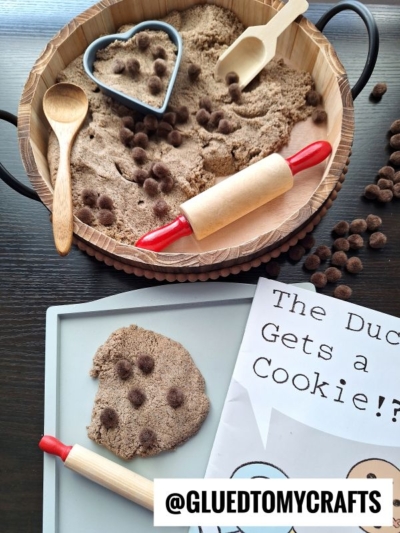 A sensory play setup with brown kinetic sand, brown pom poms, toy rolling pins, a wooden tray, a heart-shaped cookie cutter, and a storybook titled The Duck Gets a Cookie?! @GLUEDTOMYCRAFTS.