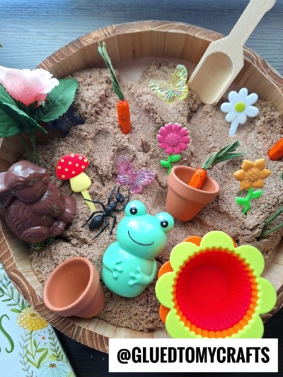 A sensory bin filled with sand, a toy frog, a chocolate bunny, plastic flowers, carrots, ants, butterflies, a wooden scoop, and small flowerpots. The scene is colorful and playful. @GLUEDTOMYCRAFTS is written at the bottom.