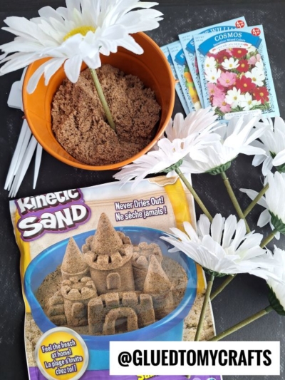 A package of Kinetic Sand, a bowl of sand, white artificial daisies, and flower seed packets are arranged on a black surface. A label in the bottom right corner reads “@GLUEDTOMYCRAFTS.”.