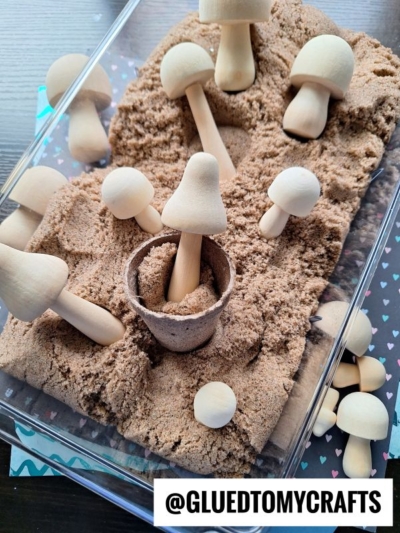 A clear plastic container filled with brown kinetic sand and several unfinished wooden mushrooms. One mushroom stands upright in a small brown pot. The container sits on a dark surface with a heart-patterned cloth underneath.
