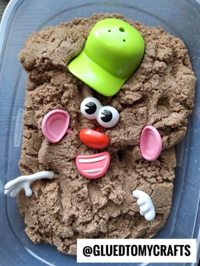 Plastic Mr. Potato Head parts—a green hat, white arms, pink ears, eyes, a nose, and a mouth—arranged on brown kinetic sand in a plastic container. @GLUEDTOMYCRAFTS is written at the bottom.