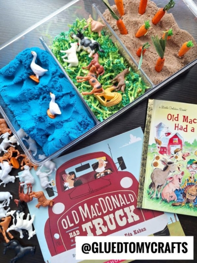 A flat lay of farm-themed children’s activities: blue and brown kinetic sand with small animal figures, green shredded paper, toy vegetables, and two Old MacDonald books. A @gluedtomycrafts label is at the bottom.