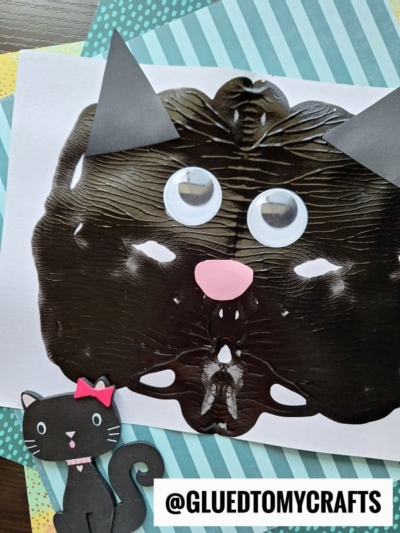 A black painted cat face made with PAINT SPLAT CRAFTS by folding paper, with googly eyes, paper ears, and a pink nose sits on layered blue and striped backgrounds. A cartoon black cat with a pink bow is in the bottom left corner.