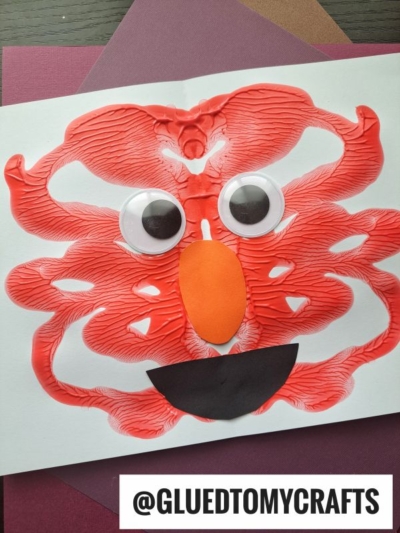 A red, abstract monster face made from PAINT SPLAT CRAFTS, with two large googly eyes, an orange oval nose, and a black smiling mouth on white paper. The handle @GLUEDTOMYCRAFTS is at the bottom.