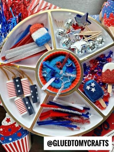 A round tray holds patriotic craft supplies like red, white, and blue pom-poms, pipe cleaners, popsicle sticks, paper rockets, star sequins, and glittery decorations. The image is labeled @GLUEDTOMYCRAFTS.