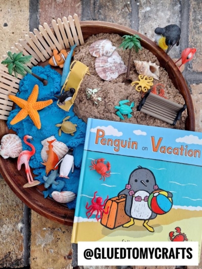 A children’s book titled Penguin on Vacation sits beside a round tray filled with blue and brown kinetic sand play ideas—sensory sand, shells, toy animals, and beach-themed decorations. A watermark reads @GLUEDTOMYCRAFTS.