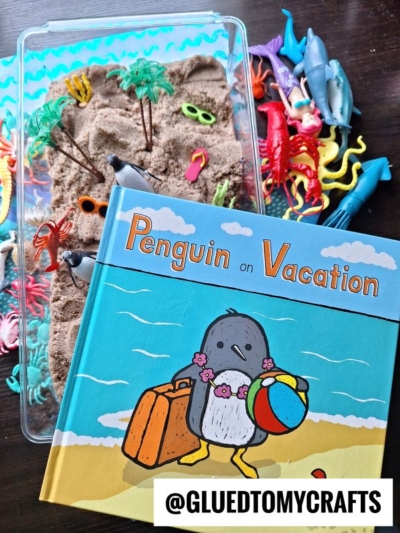 A children’s book titled Penguin on Vacation is placed on a table next to a tray of kinetic sand with toy palm trees, sunglasses, and various plastic sea animals scattered around.