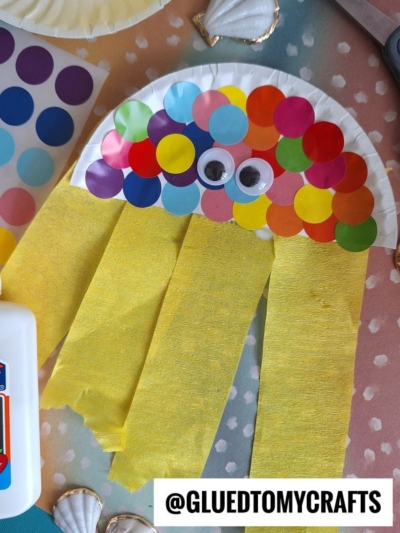 A colorful jellyfish craft made from a paper plate with multicolored dot stickers, googly eyes, and yellow crepe paper tentacles on a polka-dot background. @GLUEDTOMYCRAFTS is written at the bottom.