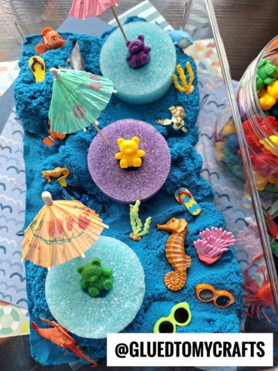 A sensory bin filled with blue kinetic sand, foam circles, toy bears, mini umbrellas, sea animal figurines, flip-flop charms, and colorful coral, arranged to look like a beach scene. @GLUEDTOMYCRAFTS is written at the bottom.