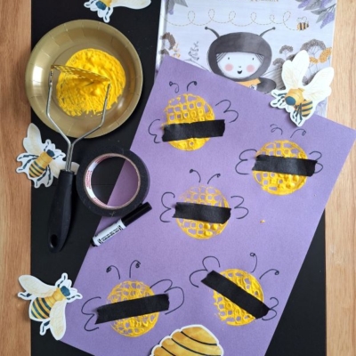 A childrens craft setup shows a book titled The Little Bee, yellow paint, a potato masher, black tape, and a sheet with bee and honeycomb art, surrounded by cut-out bees. @GLUEDTOMYCRAFTS is displayed at the bottom.