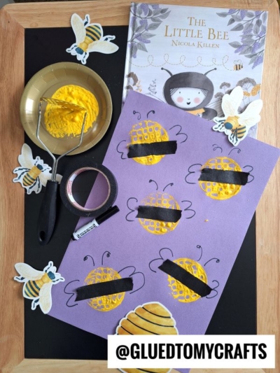 A childrens craft setup shows a book titled The Little Bee, yellow paint, a potato masher, black tape, and a sheet with bee and honeycomb art, surrounded by cut-out bees. @GLUEDTOMYCRAFTS is displayed at the bottom.