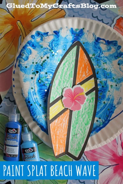 A colorful beach craft featuring a paper plate painted with blue swirls and glitter to resemble ocean waves, topped with a paper surfboard decorated with orange, green, and pink shapes. Two blue paint bottles are nearby.