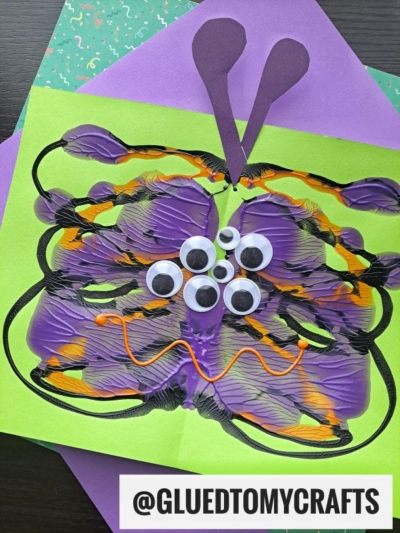 A colorful PAINT SPLAT CRAFTS butterfly made from purple, black, and yellow paint, decorated with googly eyes and orange pipe cleaner, sits on green and purple paper. @GLUETOMYCRAFTS is printed at the bottom.
