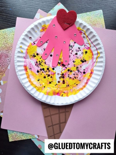 A colorful PAINT SPLAT CRAFTS ice cream cone, made from a paper plate with marbled paint, topped with a pink paper handprint and red heart, is displayed on pastel paper. The handle @GLUEDTOMYCRAFTS appears at the bottom.