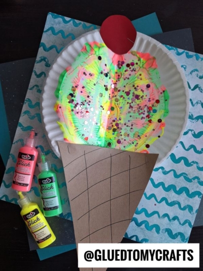 A paper plate craft shaped like an ice cream cone features colorful paint splat crafts and glitter as the scoop, a red circle for the cherry on top, brown paper for the cone, bottles of paint, and a “@GLUEDTOMYCRAFTS” label nearby.