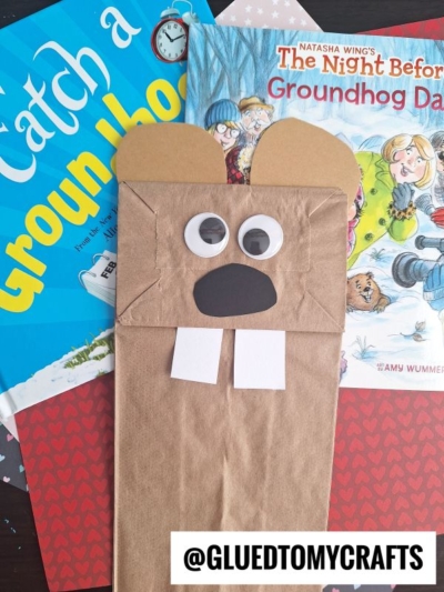 A brown paper bag puppet decorated as a groundhog with googly eyes, paper teeth, and ears sits atop two children’s books about Groundhog Day—perfect inspiration for your next PAPER BAG CRAFT IDEAS. The Instagram handle @gluedtomycrafts is visible at the bottom.