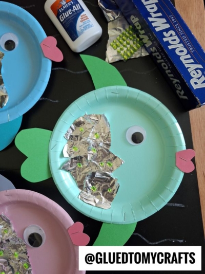 Colorful paper plate fish crafts with green fins, pink tails, a large googly eye, and foil scales with green dots. Glue and foil supplies are visible in the background. @GLUEDTOMYCRAFTS text is at the bottom.